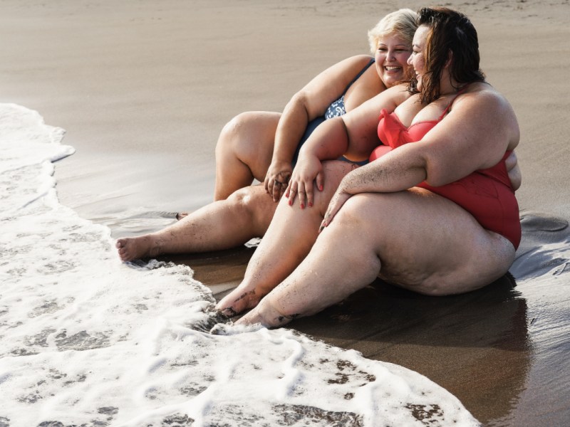 Zwei gut gelaunte, vollschlanke Frauen im Bikini sitzen am Strand am Wasser.