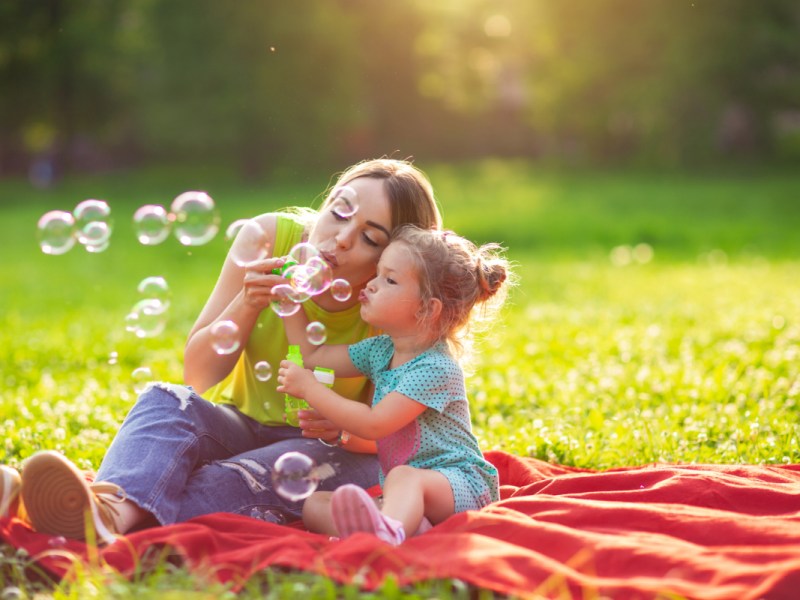 Eine Mutter sitzt mit ihrer Tochter auf einer Wiese und bläst Seifenblasen.