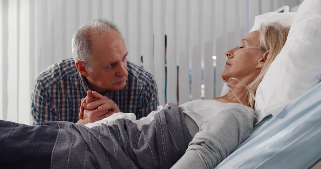 Ein älterer Mann besucht seine Frau im Krankenhaus und hält ihre Hand.