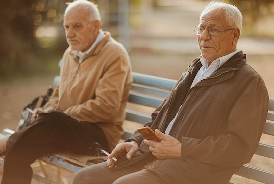 Zwei Rentner sitzen auf einer Parkbank und rauchen eine Zigarette.