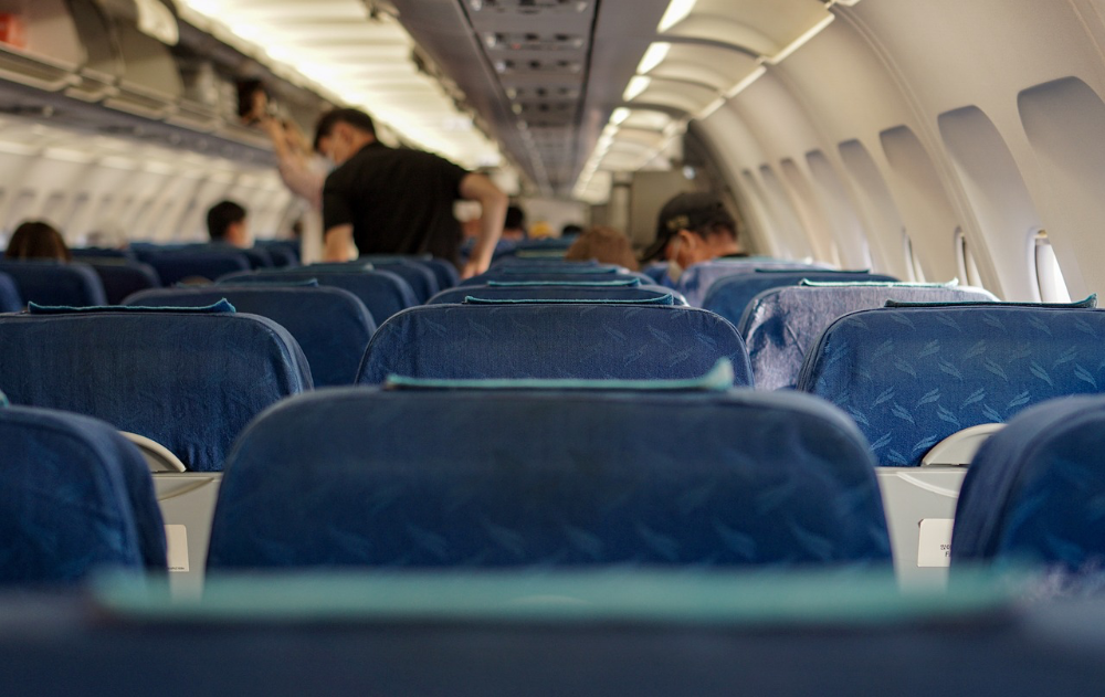 Eine Flugzeugkabine vom Sitz aus fotografiert. Einige Passagiere stehen von ihren Sitzen auf und holen ihr Gepäck aus der Gepäckablage des Flugzeugs.